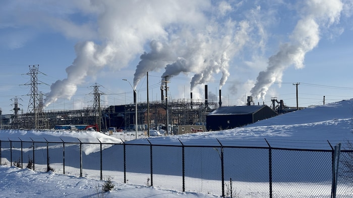 Un plan large montre un complexe industriel avec plusieurs cheminées d'où s'échappe une épaisse fumée blanche. Le paysage est enneigé au premier plan, derrière une clôture grillagée. Des pylônes électriques se dressent sur la gauche sous un ciel bleu parsemé de nuages blancs.
