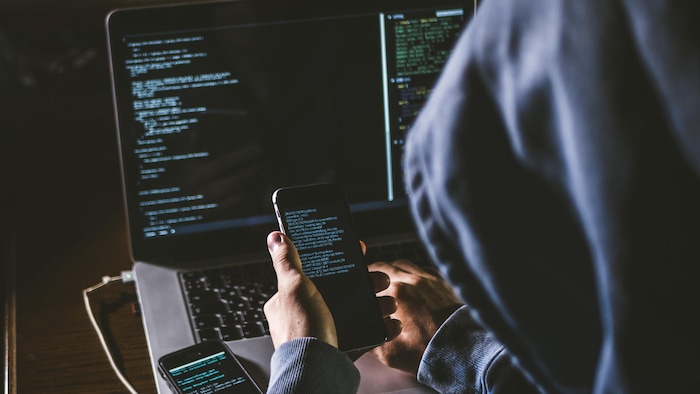 Un pirate informatique assis en face d'un ordinateur et tenant un téléphone portable.