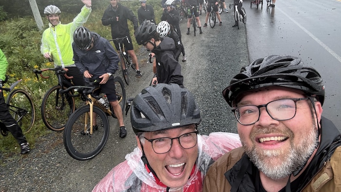 Martin Lavoie souriant à la caméra avec un autre participant, devant plusieurs autres cyclistes.