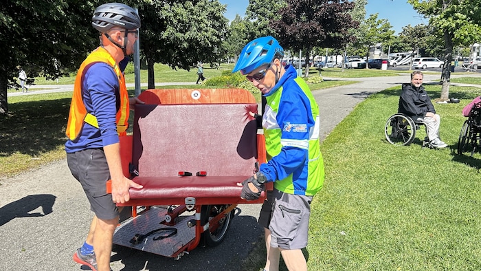 Deux hommes transportent une petite banquette pour l'installer sur un vélo adapté.