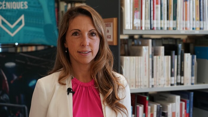 Une femme souriante avec un gilet rose et un veston blanc dans une bibliothèque.