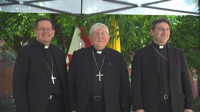 Trois hommes, portant le col romain, sourient côte à côte.