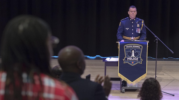 Des gens se lèvent et applaudissent le chef de police en uniforme devant un lutrin.