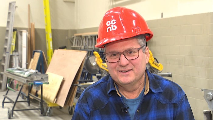 Un homme coiffé d'un casque de construction rouge décoré du logo du CCNB est assis dans un atelier.