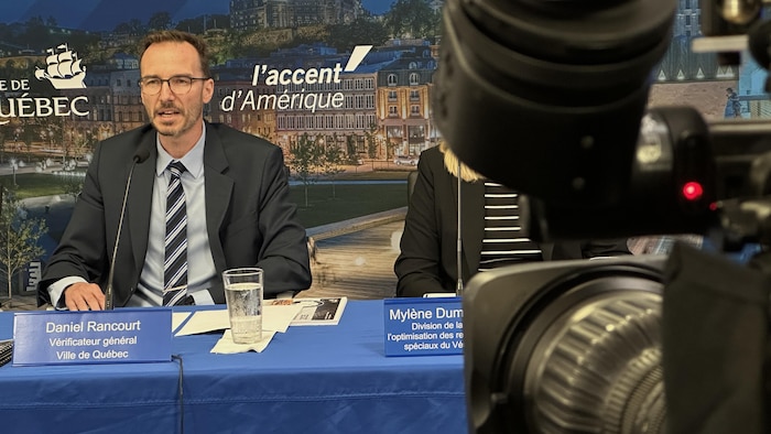 Un homme vêtu d'un veston-cravate parle au micro devant une caméra.