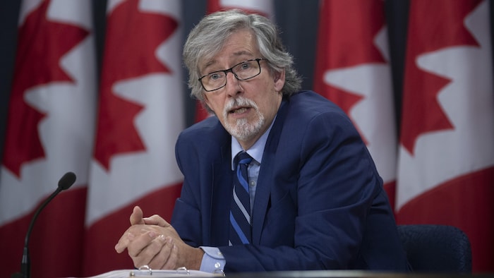 Un hombre fotografiado en primer plano frente a una fila de banderas canadienses.