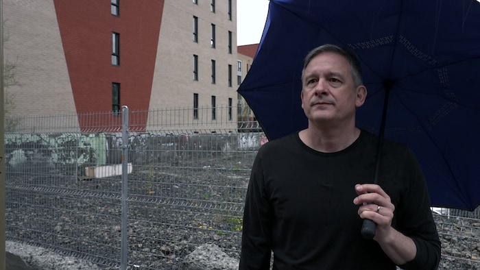 Un homme, l'air découragé, tient un parapluie devant un terrain vague.