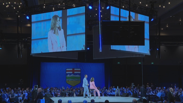 Danielle Smith assise sur un tabouret haut au centre d'une scène, dans une salle comble.