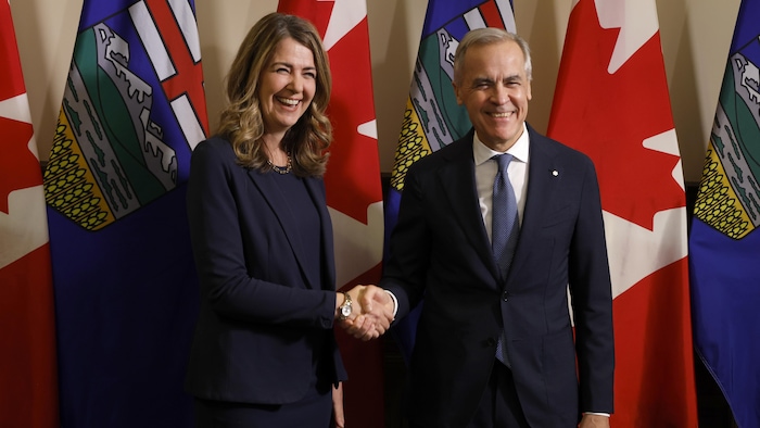 Danielle Smith et Mark Carney se serrent la main en souriant devant des drapeaux de l'Alberta et du Canada.