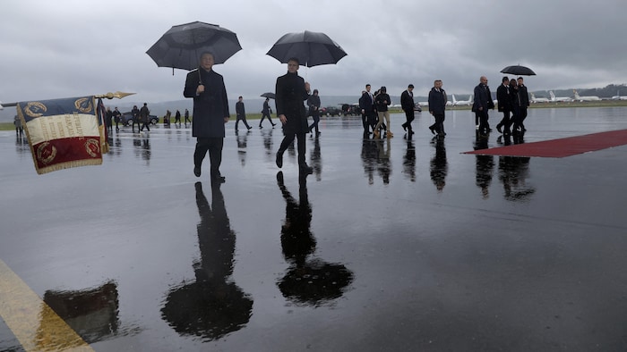 Les deux hommes marchent sur le tarmac d'un aéroport, chacun portant un parapluie.