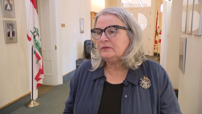 La ministre est tournée vers la gauche et parle, un micro pointé vers elle, dans un couloir de Province House.