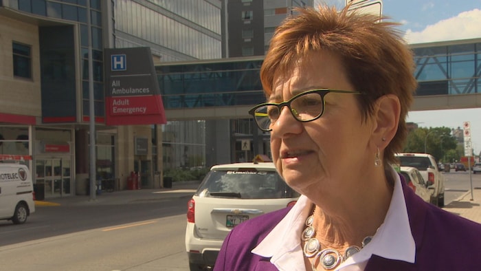 Une dame aux courts cheveux roux s'exprime dans la rue, avec en arrière plan le Health Science Center de Winnipeg.