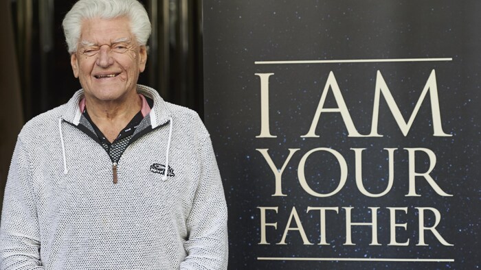 L'acteur devant une affiche où on peut lire la célèbre phrase de son personnage Darth Vader « Je suis ton père » en anglais.