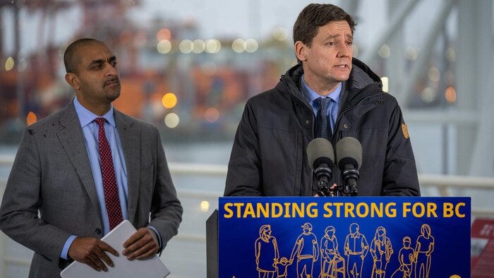 David Eby au podium devant des micros et Ravi Kahlon à gauche.