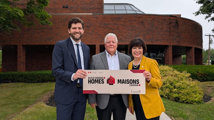 Les trois personnes devant un immeuble portent une pancarte sur laquelle il est écrit «Maisons Canada».