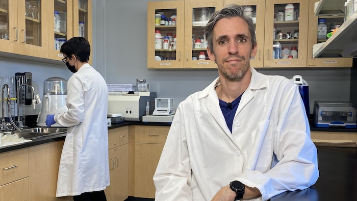 David Joly est assis dans son laboratoire, pendant qu'une étudiante manipule des outils scientifiques au comptoir derrière lui.