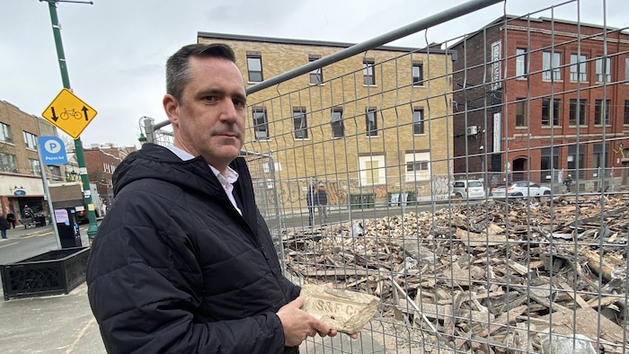 David Lacoste se tient debout devant les ruines, la brique à la main.