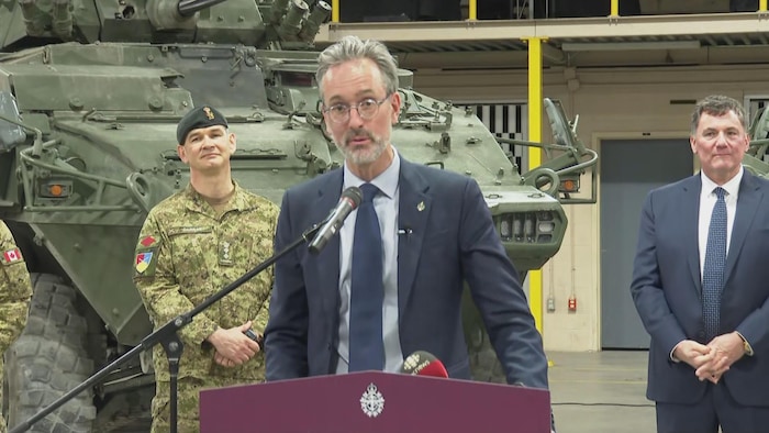 Un homme parle dans un micro en conférence de presse, des soldats et un char d'assaut derrière lui.