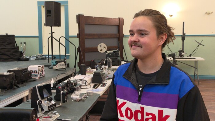 Une personne pose devant une table où se trouvent des caméras analogiques et d'autres appareils.