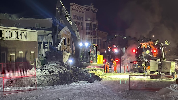 Des employés tentent de réparer une conduite d'eau dans une rue, une pelle mécanique à l'avant-plan.