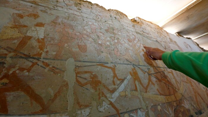 Un homme montre du doigt des peintures anciennes égyptiennes sur un pan de mur.