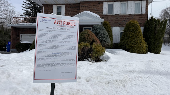 Un gros plan montre une pancarte d'avis public de la Ville de Saguenay concernant une demande de démolition. L'affiche est plantée dans la neige devant une résidence en briques, avec des conifères et un garage visibles en arrière-plan.