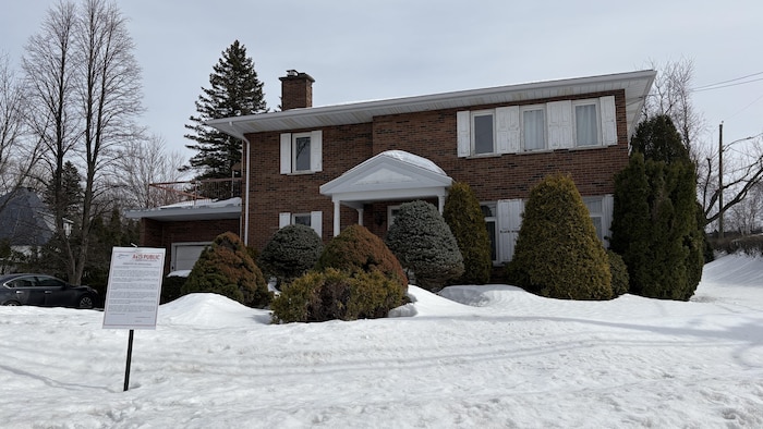Une maison à deux étages en briques brunes est entourée de neige. Plusieurs arbustes conifères bordent la façade. Au premier plan, une pancarte d'avis public est plantée dans un banc de neige sous un ciel grisâtre.