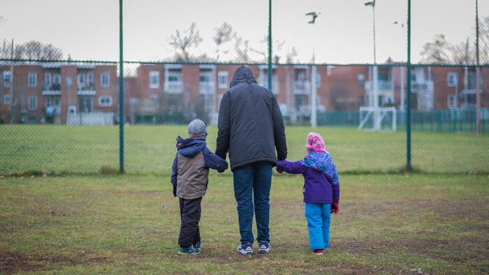 Un homme tient un garçon et une fillette par la main. Tous sont de dos et marchent vers la clôture d'un terrain de soccer.