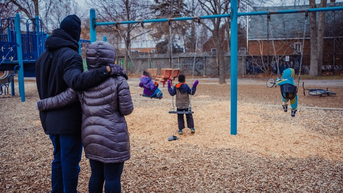 Un couple, de dos, regarde trois enfants assis dans les balançoires d'un parc.