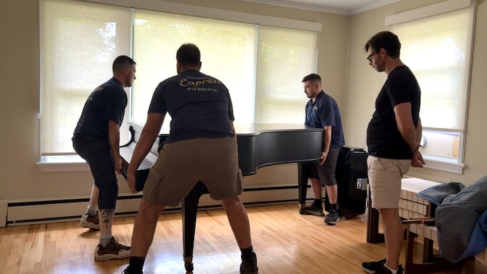 Trois hommes lèvent un piano à queue et un homme les regarde.