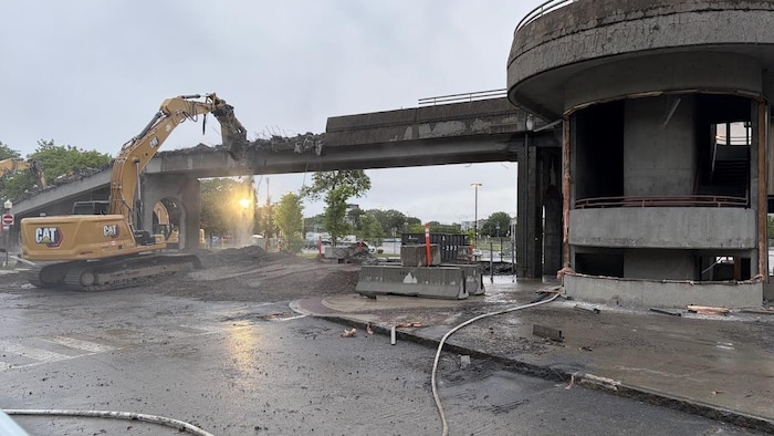 Une pelle mécanique est en train de démolir une structure de béton qui surplombe une autoroute.