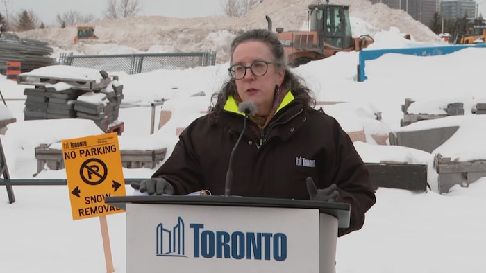 Barbara Grey qui donne une conférence en extérieur au milieu de tas de neige et à côté d'une pancarte qui interdit le stationnement.