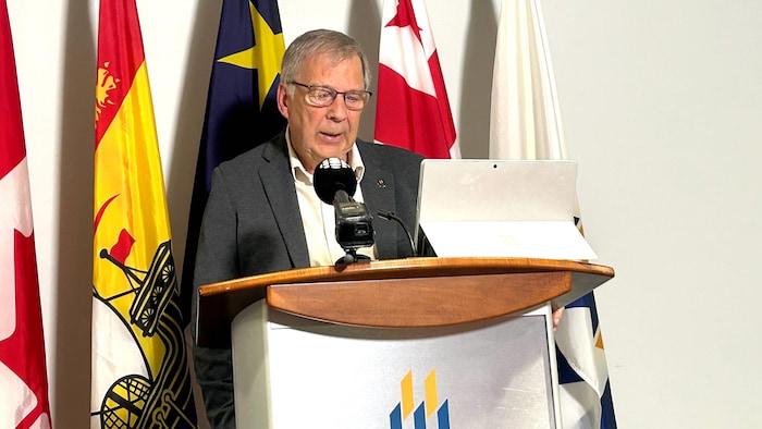 Un homme est debout à un lutrin décoré du logo de l'Université de Moncton, sur lequel est posée une tablette électronique. Il regarde l'écran et parle dans un micro.