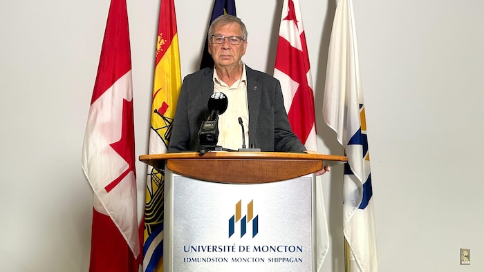 Denis Mallet debout à un lutrin décoré du logo de l'Université de Moncton. Il regarde droit devant.