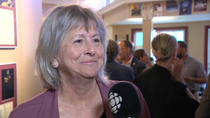 Avec beaucoup de gens derrière elle dans la salle, Denise Comeau Désautels sourit et regarde vers la droite, un micro pointé vers elle.