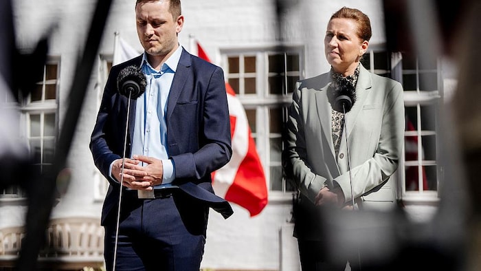 Un homme et une femme se tiennent debout à l'extérieur, devant des microphones, avec un drapeau danois en arrière-plan.
