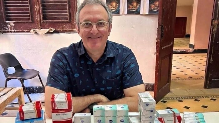 Un homme pose devant la caméra et est assis à une table sur laquelle se trouve plein de médicaments.