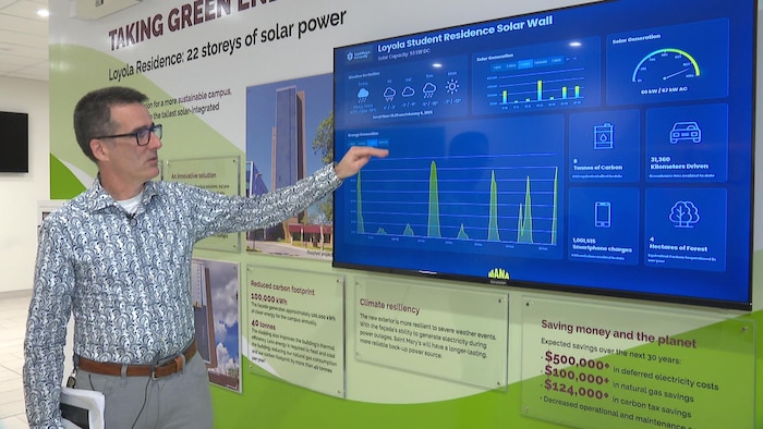 Un homme qui porte une chemise avec des motifs audacieux est debout devant un écran géant posé au mur et pointe les données qui y sont affichées au sujet de la production d'énergie solaire du bâtiment.