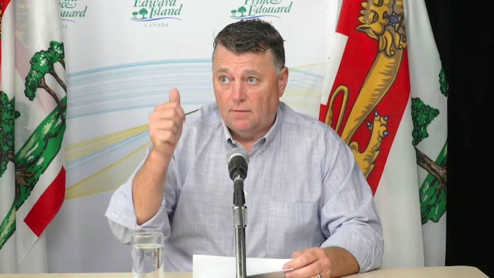 Assis à une table devant un micro et tenant des documents, Dennis King lève un pouce en l'air pour signifier qu'il est prêt à commencer son allocution.