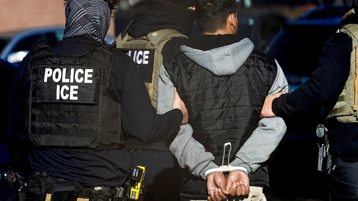 Deux policiers tiennent fermement un homme aux mains liées dans son dos.