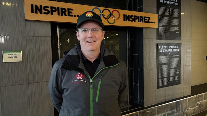 Derrick McLeod porte une casquette avec le logo du drapeau du Nouveau-Brunswick. Il a un manteau à l'effigie de Patinage de vitesse Nouveau-Brunswick. Il se tient devant une bannière des Jeux olympiques.