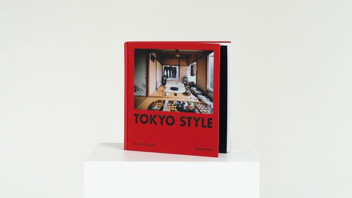 Un livre à la couverture rouge vif intitulé « TOKYO STYLE » par Kyoichi Tsuzuki, posé bien droit sur un socle blanc minimaliste. Au centre de la couverture, une photographie montre l'intérieur d'un appartement japonais authentique et très rempli, avec des tatamis au sol, des vêtements suspendus et des objets du quotidien. L'arrière-plan de l'image est entièrement blanc.