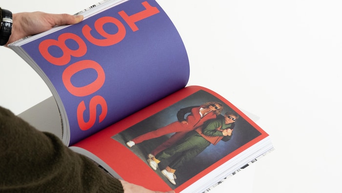 Gros plan sur les mains d'une personne feuilletant un épais livre d'art posé sur un socle blanc. La page de gauche, sur fond bleu-violet, affiche en grands caractères rouges la typographie « 1980s ». La page de droite présente, sur un fond rouge vif, une photographie rétro de deux personnes portant des lunettes de soleil et des tenues emblématiques des années 80. L'arrière-plan de la photo est entièrement blanc.