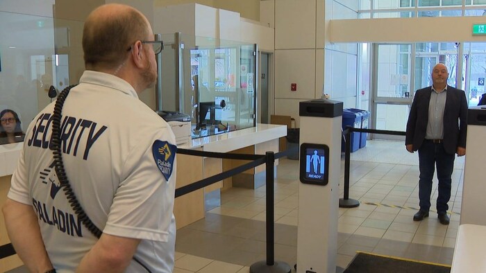 Un homme se tient debout dans le hall de l'hôpital face à un agent de sécurité. Le signal du détecteur de métal est neutre avant qu'il ne passe devant.