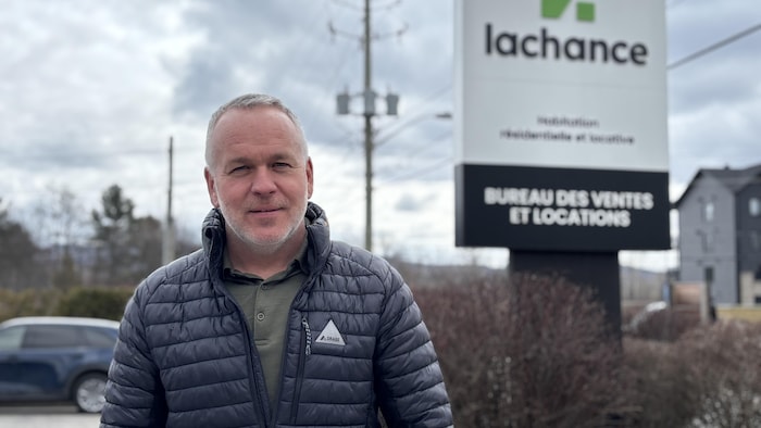 Patrick Lachance pose devant une affiche qui indique « Lachance, bureau des ventes et locations »