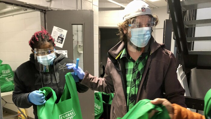 Deux personnes avec visière et couvre-visage donnent des sacs verts contenant des denrées alimentaires.