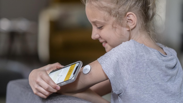 Une jeune fille place un téléphone cellulaire près d'un capteur de glycémie posé sur son bras.