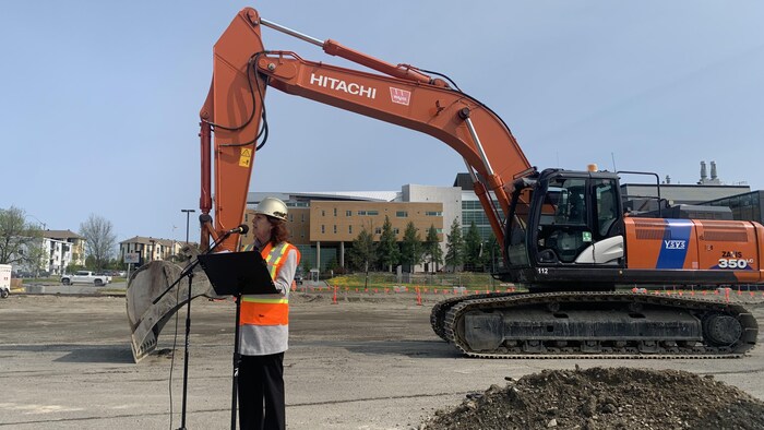 Une femme portant un casque de sécurité fait un discours dans un chantier de construction, en arrière d'elle une pelleteuse est stationnée.