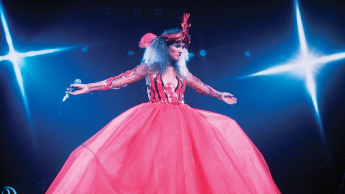 L'artiste sur scène, les bras étendus à l'horizontale, portant une grande robe bouffante à partir de la taille. 