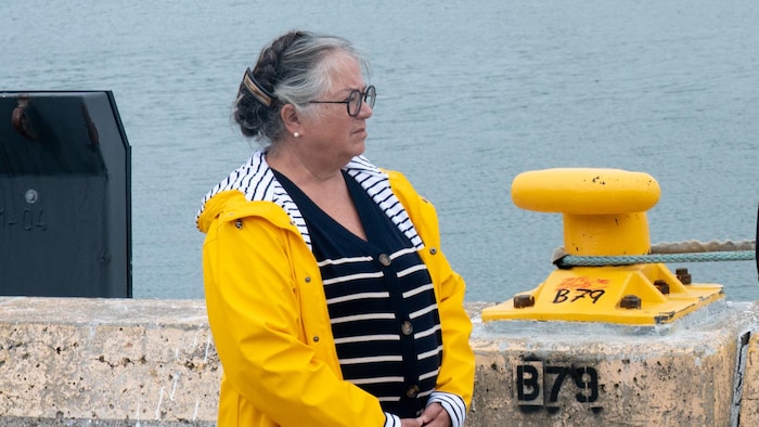 Diane Lebouthillier de profil, portant un imperméable jaune, debout les mains croisées sur un quai, près de l'eau et devant un bollard peint en jaune.